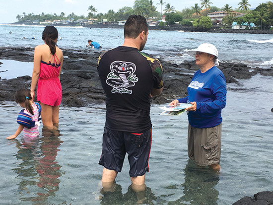 ReefTeach volunteer Mary Saarloos wades into the water to talk to visitors about reef etiquette, using laminated photos with symbols to communicate in any language. Kahalu‘u Bay is the most popular swimming site in Kona, attracting 400,000 visitors a year.