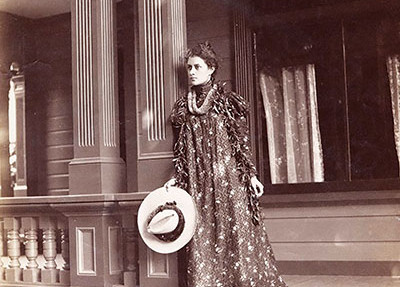 Princess Ka‘iulani standing on top of steps on the porch of her house at ‘Āinahau; wearing the holokū and lei, 1898. public domain photo by Frank Davey