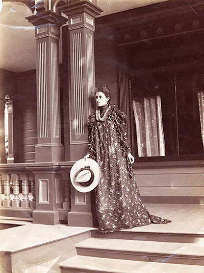 Princess Ka‘iulani standing on top of steps on the porch of her house at ‘Āinahau; wearing the holokū and lei, 1898. public domain photo by Frank Davey