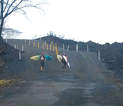 Surfers make the three-plus mile trek with their boards to Pohoiki breaks. Prior to the freshly graded lava-covered road, many carried surfboards over an ‘a`ā trail along the shoreline. photo by Marcia Timboy